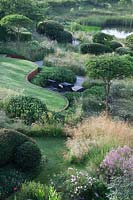 Overview of garden with countryside views beyond. Curving rusted Corten steel walls, Pinus mugo, clipped Crataegus x lavalleei 'Carrierei', Taxus baccata - Yew topiary, meadows and  natural pond. Borders with Stipa gigantea, Cenolophium denudatum, Eryngium giganteum, Thalictrum Elin and Rosa 'Rosemoor'.