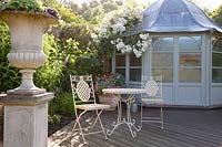 Decorative metal furniture on decked patio with stone urn and summerhouse with Rosa