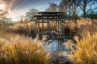 View across reflective pool and swathes of Miscanthus and other tall grasses to seating area within modern oak summerhouse in the minimalist front garden in Winter at Bury Court Gardens, Hampshire. Designed by Christopher Bradley-Hole.