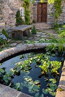 Seating area in Spanish style garden with Hydrangea macrophylla 'Forever Blue' in container and pond with waterlilies - The Pazo's Secret Garden. RHS Hampton Court Flower Show, 2017 - Designer: Rosie McMonigall. Sponsor: Turismo de Galicia, North Spain.
