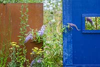 Corten steel structures designed by sculptor Simon Probyn with Buddleja davidii 'Wisteria Lane' and perennials including Achillea 'Moonshine' and Pilosella aurantiaca with blue wooden wall - Brownfield Metamorphosis. RHS Hampton Court Palace Flower Show 2017 - Designer: Martyn Wilson. Sponsor: St. Modwen Properties PLC