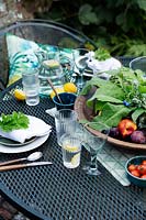 Outdoor dining area under a fig tree on a brick patio. Table laid with greens and fresh whites, napkins dressed with a geranium leaf and the centre of the table has a wooden bowl of fresh green leaves from the garden surrounded by fruit