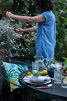Dusk falls on the outdoor dining area under a fig tree. Woman puts fairy lights in the trees to create a cosy setting and light up the area. Table laid with greens and fresh whites and napkins dressed with a geranium leaf