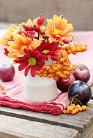 Autumnal display with Chrysanthemums in reds, yellows and oranges, rosehips and orange Pyracantha berries on a table with plums