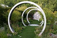 Breast Cancer Now Garden: Through the Microscope -Rings representing microscope lens with Acer trees and soft planting of Briza media, Geum 'Totally Tangerine', Gem 'Mai Tai' and Iris 'Natchez Trace' - RHS Chelsea Flower Show 2017