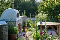 Outdoor pizza oven and cow parsley in a jug on a wooden table - The Refuge Garden in aid of Help Refugees UK, RHS Malvern Spring Festival 2017 