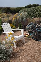 Seaside themed front garden with white Adirondack chair, planted with coastal plants  including Cineraria 'Silver Dust', Santolina 'Lambrook Silver' , Stipa tenuissima, purple Heuchera and decorated with driftwood sculptures, lobster pots, fishing floats. Behind is an Elaeagnus x ebbingei  hedge and sea view.