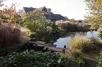 View of the house from The middle pond with Japanese zig-zag bridge. Viburnum davidii, Gunnera, Miscanthus, Pennesetum alopecuroides and Prunus foliage - Brightling Down Farm