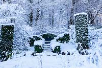 The Wild Garden. Columns of Yew - Taxus baccata. Hedges of Box - Buxus sempervirens surrounding seat. View to the wood behind. Garden in snow. Veddw House Garden, Monmouthshire, Wales, UK. The garden was created since 1987 by garden writer Anne Wareham and her husband, photographer Charles Hawes. The garden opens to regularly to the public in the summer months.