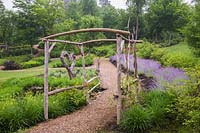 Mulch footpath through rustic wooden arbour edged with Nepeta and Hemerocallis 'Stella de Oro'in early summer. Le Jardin de Francois garden, Quebec, Canada. 