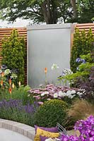 A stainless steel sheet water wall cascade flanked by Taxus baccata 'Fastigiata Aureomarginata' and  Kniphofia 'Royal Standard', Achillea 'Lilac beauty' and Leucanthemum x superbum 'Aglaia' in front in the 'Facing Fear, Finding Hope' Garden at Tatton RHS Flower Show 2017