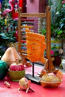A collection of eclectic brightly coloured ornaments on a red retro laminex table underneath a covered outdoor entertaining area featuring metal fruit, an childs abacus and an Thai style head ornament painted orange.