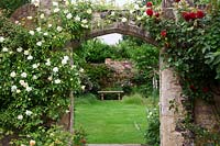 Stone and flint archway covered with Rosa 'Alberic Barbier' and Rosa  'Dublin Bay'. Rosa 'Madame Isaac Pereire' beyond arch on wall above stone bench.