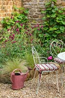 Small courtyard garden with stone wall, chairs and watering can, Stipa tenuissima,  Cirsium rivulare 'Atropurpureum', Vitis coignetiae
