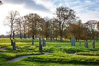 Stone circle comprising a winding labyrinth with monoliths of slate set in grass, backdrop of deciduous trees in autumn
