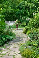 Garden path with shrubs and perennials, garden bench and planted iron urn
