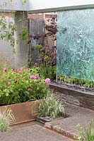 The Silent Pool Gin Garden, view of sunken garden with glass wall water feature, corrugated metal fencing, recycled concrete pillars, clay brick wall and paving, roses, grasses on  copper raised bed – Designer: David Neale - Sponsor: Silent Pool Gin