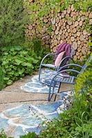 A pair of metal seats infront of a wall made from logs. The Art of Viking Garden at RHS Chelsea Flower Show 2019. Design: Paul Hervey-Brookes. Sponsor: Viking Cruises.