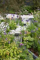 The Manchester Garden, view of sculptures and cottage garden planting, with hexagonal stepping stones over the pond, bordered by Rodgersia, Iris sibirica 'Persimmon, Iris sibirica 'Caeser's Brother', and Iris sibirica 'Harpswell Happiness' - RHS Chelsea Flower Show 2019 - Designer: Exterior Architecture, exteriorarchitecture.com 
