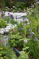 The Manchester Garden, view of sculptures and cottage garden planting, with hexagonal stepping stones over the pond, bordered by centranthus ruber 'Albus', rodgersia, and iris sibirica - Designer: Exterior Architecture, exteriorarchitecture.com. RHS Chelsea Flower Shwo 2019
