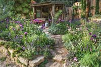 Stone steps and path leading to a wooden lean to shelter in a Mediterranean style planted garden. Stone walled raised beds with purple and pink flowering plants and silver foliage plants. The Donkey Sanctuary: Donkeys Matter. Design: Christina Williams and Annie Prebensen. Sponsor: The Donkey Sanctuary. RHS Chelsea Flower Show 2019
