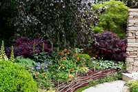 Miles Stone: The Kingston Maurward Garden. Colourful woven screen fencing a raised bed by curved path. Designer: Michelle Brown, Sponsor: Miles Stone. Kingston Maurward College. RHS Chelsea Flower Show 2019