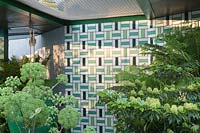 Glazed tiled wall in shaded area of garden, planted with Angelica and ferns. The Greenfingers Charity Garden. Designed by Kate Gould Gardens, sponsored by Greenfingers Charity, RHS Chelsea Flower Show, 2019.
