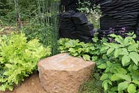 The M and G Garden, view of green planted woodland garden with shade loving plants English ironstone stone seat, charred oak walls sculpture by Johnny Woodford – Designer: Andy Sturgeon - Sponsor: M and G 