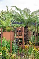 Urban garden with Dicksonia antartica, Crocosmia 'George Davison' and Veronicastrum virginicum 'Album', Corten water containers and reclaimed, rusted drainage pipes, surrounded by burnt effect fencing - Bee's Gardens, The Penumbra, RHS Tatton Park Flower Show 2018