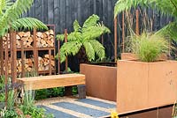 Slate paving, gravel, oak bench, Corten water container, Dicksonia antarctica and log store surrounded by a burnt effect fence - Bee's Gardens: The Penumbra, RHS Tatton Park Flower Show 2018