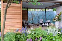 Armchairs and table in timber pavilion with eddy art wall and planting of Dodecatheon meadia, Euphorbia characias subsp. wulfenii 'White Swan' and Camassia leichtlinii 'Maybelle' - The Morgan Stanley Garden for the NSPCC - Sponsor: Morgan Stanley - RHS Chelsea Flower Show 2018