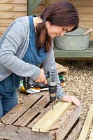 Woman using electric drill to drill holes in lengths of wood. 
