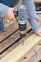 Woman using electric drill to drill holes in lengths of wood. 