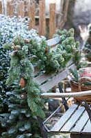 A bench inside a garden powdered by snow. The bench is decorated with a festoon 
out of fir tree and a basket filled with caucasian fir, 4 red candles and Pinus 
strobus cones. You see a quick advent decoration or wreath