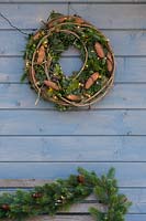 A wreath hanging on the wall of blue summer house featuring fairy lights, 
mixed greenery, and pine cones 