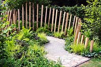 Lush green planting along path of crazy paving and crushed sandstone, with timber posts along one edge. Calm In Chaos Garden, Sponsored by Groundwork UBS Wealth Management, RHS Tatton Park Flower Show, 2018.  