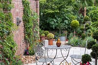 An enclosed patio seating area with small fountain inbetween nectarines and figs fan-trained up a sunny brick wall.  On the right, cloud pruned conifers are underplanted with colourful perennials.