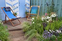 Blue and white beach huts and deck chairs bordered by blue and white coastal planting. Southend Council 'By The Sea' garden at RHS Hampton Court Flower Show, London, 2017.