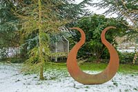 Rusted steel Lyre sculpture set in wild garden with conifers and evergreens. 