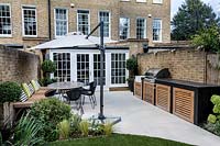 Contemporary back garden with white stone patio, with garden furniture, parasol
 and wooden seating dressed with garden cushions. 
To the right is a garden kitchen and dining area with built-in barbecue.
 In the forground  is an artificial lawn and a bed with Stipa tenuissima
 and Ilex crenata clipped to a ball