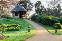 Path leading up to the Swiss Cottage with thatched roof, sloping lawn
 with daffodils and shrubs

