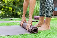 Woman standing barefoot in garden, rolling up yoga mat. 
