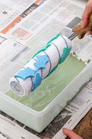 Woman lowering roller with foam patterns into tupperware of green paint.