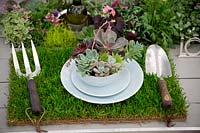 Garden table setting, with grass table mat, garden fork and trowel and succulent filled bowl. 
