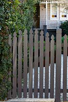 A closed, grey-painted Gothic picket gate with view of weatherboard Victorian cottage behind. 