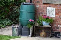 Water butt on stand by side of house with watering can, decorative crate, container plantings and wall plaque

 and planters
