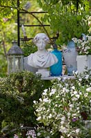 View of table of collectable in a garden, items include a woman's bust 
surrounded by lantern, jug of honesty and potted plant