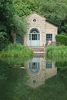 View across water and marginal planting to The Shell House Folly, reflected in the water
