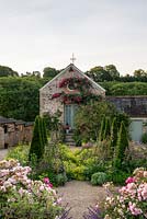 View of a stone cottage looking from the kitchen garden down the path
 of tightly clipped Taxus - Yew - pyramids with informal herbaceous planting between