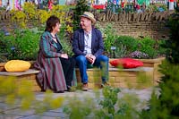 Part of the Health and Wellbeing permanent garden, Jekka McVicar and 
Joe Swift sit in conversation on brightly coloured cushions in the 
central circular sitting area, with herbs in the foreground 
